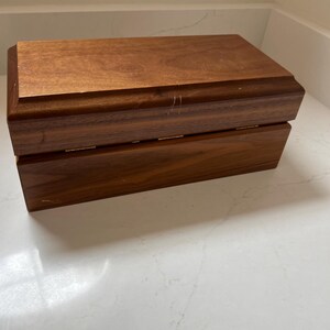 May include: A rectangular wooden box with a hinged lid. The box is made of a dark brown wood with visible grain and a slightly glossy finish. The box appears to be empty and is sitting on a white surface.
