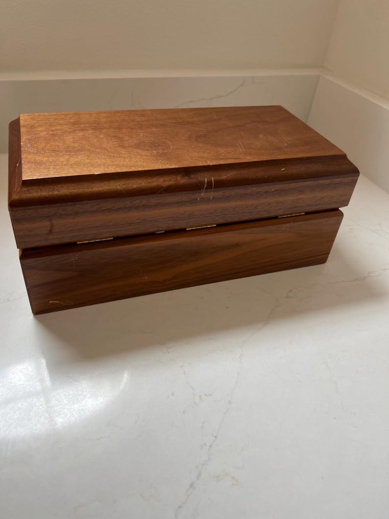 May include: A rectangular wooden box with a hinged lid. The box is made of a dark brown wood with visible grain and a slightly glossy finish. The box appears to be empty and is sitting on a white surface.