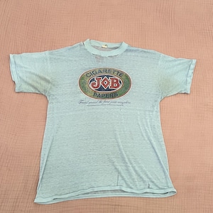 May include: Light blue t-shirt featuring an oval graphic with the words "CIGARETTE JOB PAPERS" in a vintage design. The shirt has short sleeves and a crew neck. The text "Found around the finest joints everywhere" is below the oval graphic.