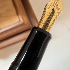 May include: Close-up of a black fountain pen with a gold nib. The nib is engraved with the brand name "NAMIKI" and other markings. The pen has a glossy black barrel and a gold-colored nib.