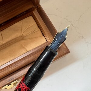 May include: A black fountain pen with a gold nib and a decorative red maple leaf design. The pen is displayed in an open wooden box with a tan interior. The pen's nib is a dark blue color.