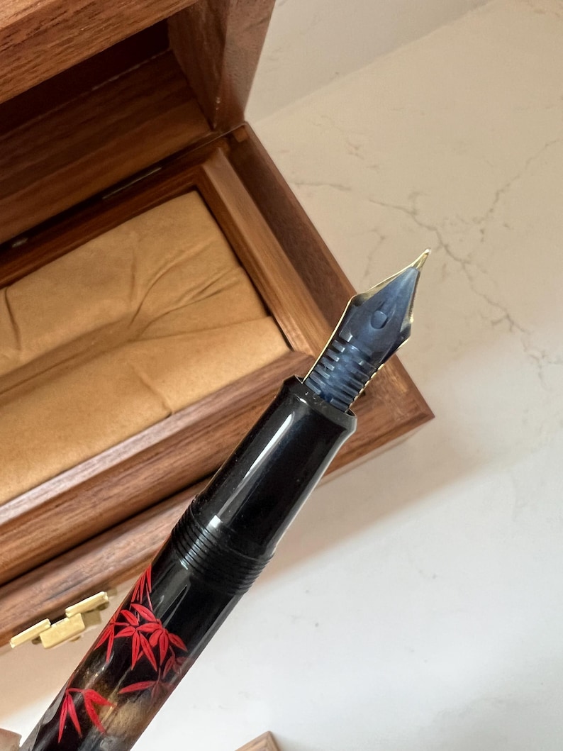 May include: A black fountain pen with a gold nib and a decorative red maple leaf design. The pen is displayed in an open wooden box with a tan interior. The pen's nib is a dark blue color.