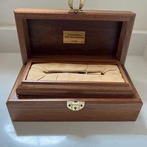 May include: A wooden box with a hinged lid, open to reveal a pen nestled in a tan-colored interior. The box is made of dark wood and has a brass clasp. A small plaque inside reads "Limited Edition White Tiger Fountain Pen 191/300."