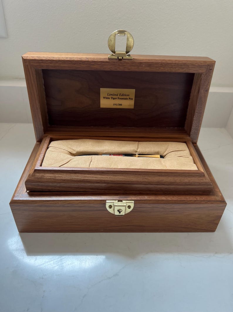 May include: A wooden box with a hinged lid, open to reveal a pen nestled in a tan-colored interior. The box is made of dark wood and has a brass clasp. A small plaque inside reads "Limited Edition White Tiger Fountain Pen 191/300."