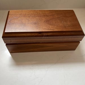 May include: A rectangular wooden box with a hinged lid. The box is made of a medium-toned wood with a smooth, polished finish. The box is sitting on a white surface.