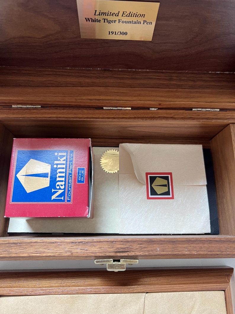 May include: A wooden box with a brass clasp, containing a red and blue Namiki fountain pen ink box, a cream-colored envelope with a gold seal, and a gold plaque that reads "Limited Edition White Tiger Fountain Pen 191/300."
