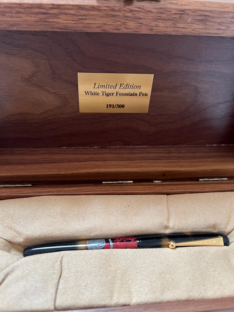 May include: A black and gold fountain pen with red and white accents, presented in a wooden box. The pen is labeled "White Tiger Fountain Pen" and is numbered 191/300.