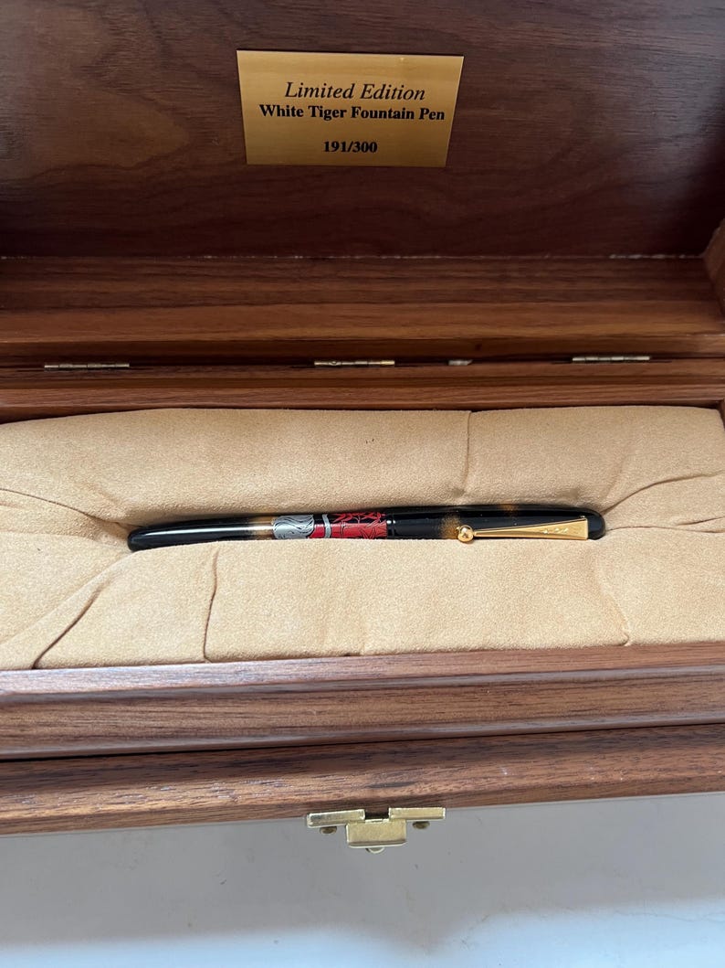 May include: A limited edition White Tiger fountain pen, number 191 of 300, displayed in an open wooden box with a tan interior. The pen is black with gold accents and a detailed design.