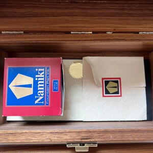 May include: Inside a wooden box, a red box of Namiki Fountain Pen Ink is visible, with a blue and gold logo. Next to it is a cream-colored envelope with a gold seal and a black and red logo. The box is open.