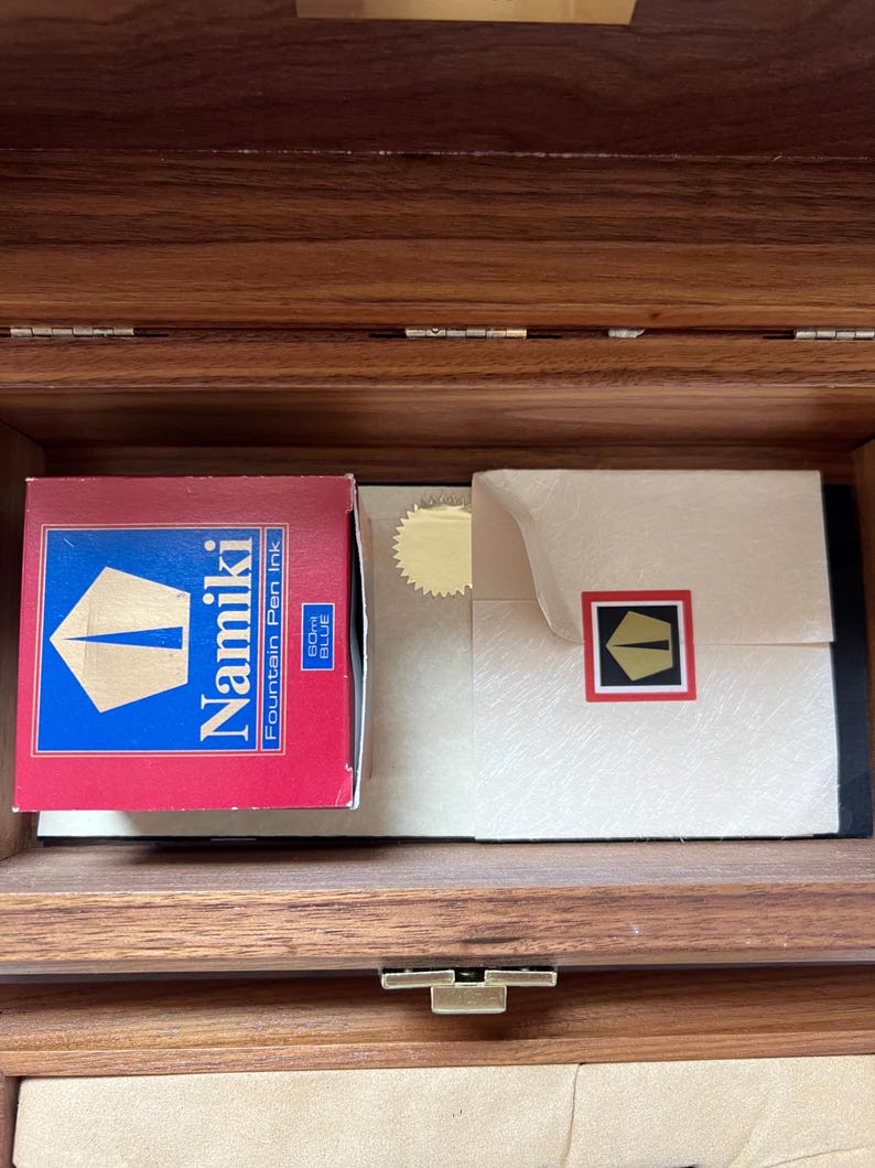 May include: Inside a wooden box, a red box of Namiki Fountain Pen Ink is visible, with a blue and gold logo. Next to it is a cream-colored envelope with a gold seal and a black and red logo. The box is open.