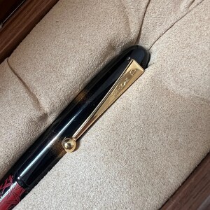 May include: A black and red fountain pen with gold-colored accents, including a clip and a small sphere. The pen is presented in a wooden box with a soft, beige interior. The pen's design features a combination of black and red.