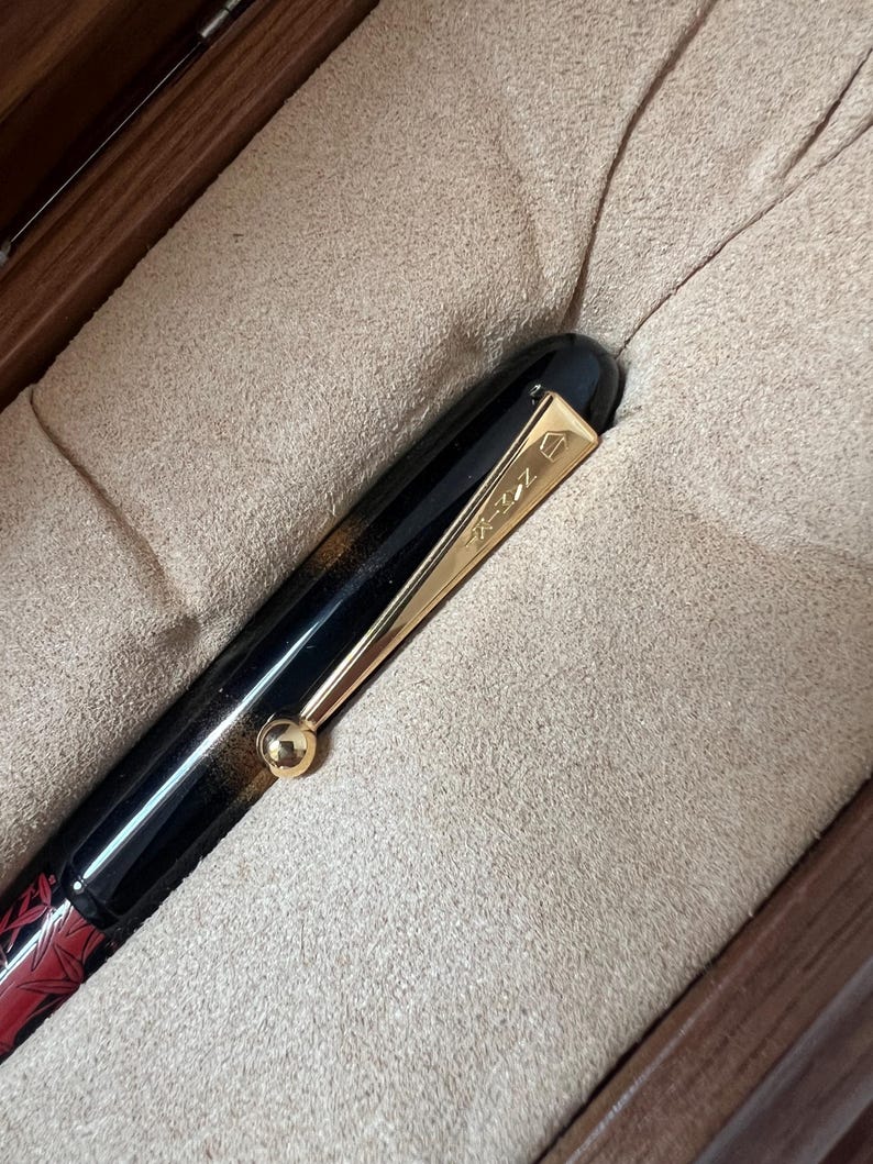 May include: A black and red fountain pen with gold-colored accents, including a clip and a small sphere. The pen is presented in a wooden box with a soft, beige interior. The pen's design features a combination of black and red.