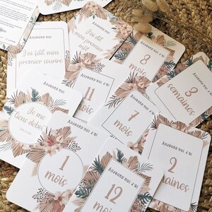 May include: A set of 12 milestone cards with a tropical theme. The cards are white with brown and green floral designs. The cards feature numbers and words in a light brown color. The cards are arranged on a woven straw surface. The text on the cards reads: 'Aujourd'hui j'ai', '4 mois', '10', 'Aujourd'hui', 'J'ai dit mon premier mot', 'Aujourd'hui j'ai', '11 mois', 'Aujourd'hui j'ai', '8 mois', 'Aujourd'hui j'ai', '7 mois', 'Aujourd'hui j'ai', '3 semaines', 'Aujourd'hui j'ai', '2 semaines', '1 mois', 'Aujourd'hui j'ai', '12 mois', 'J'ai fait mon premier sourire', 'Je me tiens debout', 'Date de l'événement'. 