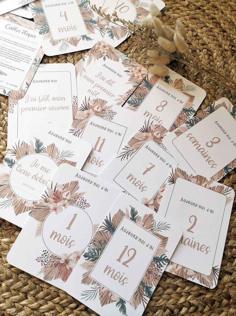 May include: A set of 12 milestone cards with a tropical theme. The cards are white with brown and green floral designs. The cards feature numbers and words in a light brown color. The cards are arranged on a woven straw surface. The text on the cards reads: 'Aujourd'hui j'ai', '4 mois', '10', 'Aujourd'hui', 'J'ai dit mon premier mot', 'Aujourd'hui j'ai', '11 mois', 'Aujourd'hui j'ai', '8 mois', 'Aujourd'hui j'ai', '7 mois', 'Aujourd'hui j'ai', '3 semaines', 'Aujourd'hui j'ai', '2 semaines', '1 mois', 'Aujourd'hui j'ai', '12 mois', 'J'ai fait mon premier sourire', 'Je me tiens debout', 'Date de l'événement'. 