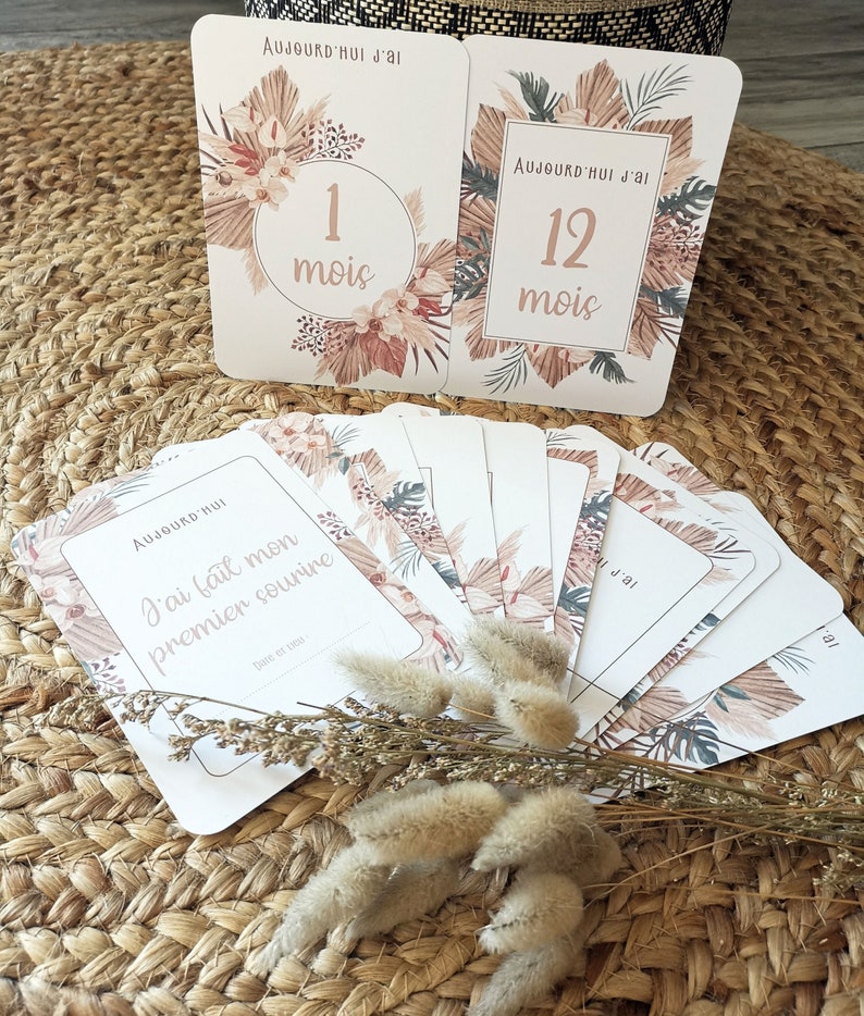 May include: A set of twelve cards with a tropical theme. The cards are white with brown and green floral designs. The cards are numbered 1 through 12 and have the text 'Aujourd'hui j'ai' and 'mois' on them. The cards also include a card that says 'Aujourd'hui' and 'J'ai fait mon premier sourire' with a space for a date.