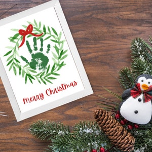 Puede incluir: Un marco blanco con una corona verde y un lazo rojo. Una huella de mano est&aacute; en el centro de la corona. El texto "merry Christmas" est&aacute; escrito en rojo debajo de la corona.