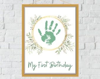 Puede incluir: Un marco dorado con fondo blanco y una huella de mano verde dentro de un círculo dorado con hojas verdes alrededor. El texto "My First Birthday" está en la parte inferior del marco.