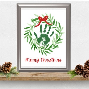 Puede incluir: Una impresi&oacute;n enmarcada con una huella de mano verde dentro de una corona verde con un lazo rojo. El texto "Merry Christmas" est&aacute; impreso debajo de la corona.
