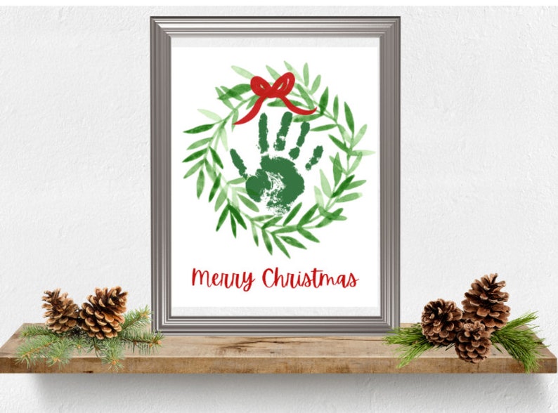 Puede incluir: Una impresi&oacute;n enmarcada con una huella de mano verde dentro de una corona verde con un lazo rojo. El texto "Merry Christmas" est&aacute; impreso debajo de la corona.