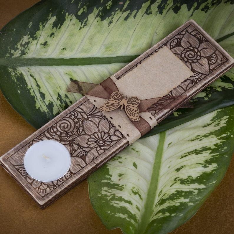May include: A wooden candle holder with a floral design and a small tea light candle. A brown ribbon with a butterfly charm is tied around the holder.