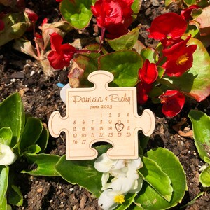May include: A wooden puzzle piece with the names "Patricia & Ricky" and the date "June 2023" engraved on it. The calendar highlights the 17th with a heart symbol. The puzzle is surrounded by red flowers and green foliage.