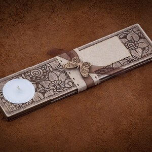 May include: A wooden candle holder with a decorative floral design and a small white candle. The holder has a ribbon tied around it with a butterfly charm.