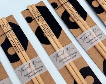 Könnte beinhalten: Ein Set brauner Holzstäbchen mit einem Dankeschön-Etikett, auf dem "Danke Stephenie & Courtney November 2023" steht. Die Stäbchen sind in braunes Papier mit einem schwarz-weißen Yin-Yang-Symbol und dem Text "ANDREW & LAN - AUGUST 17, 2024" eingewickelt.