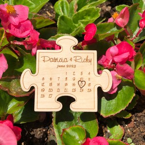 May include: A wooden puzzle piece with the names "Patricia & Ricky" and the date "June 2023" engraved on it. The calendar highlights the 17th with a heart. The puzzle is surrounded by pink flowers and green leaves.