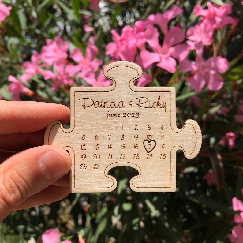 May include: A wooden puzzle piece engraved with the names "Patricia & Ricky" and the date "June 2023." The calendar highlights the 17th with a heart symbol. The puzzle is held against a backdrop of pink flowers.