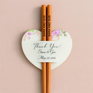 May include: Two orange pencils with "SARA AND GUS" printed on them are held by a heart-shaped card. The card has a floral design and the text "Thank You! Sara & Gus May 24, 2026". The background is a light pink.
