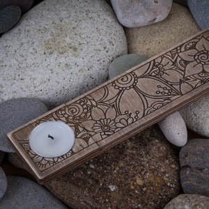 May include: A wooden candle holder with an intricate floral design. The holder has a circular cutout for a candle and is surrounded by gray stones.