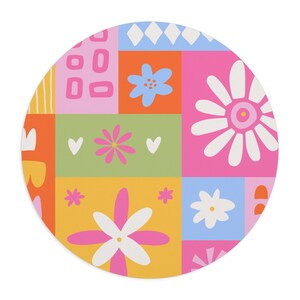 May include: A circular mouse mat featuring a vibrant patchwork design of various colourful blocks. The pattern includes illustrations of white, pink, and blue flowers, small hearts, and geometric shapes in shades of orange, yellow, pink, and blue.