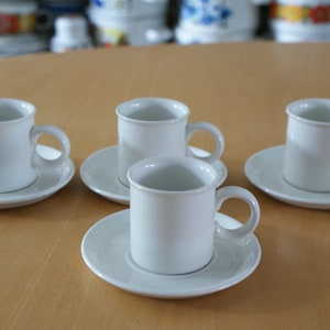 Może przedstawiać: Cztery białe ceramiczne filiżanki do kawy z pasującymi spodkami ułożone na jasnobrązowej drewnianej powierzchni. Każda filiżanka ma zaokrąglony uchwyt. Filiżanki i spodki są w jednolitym, matowym białym kolorze.