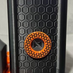 Peut inclure: Un appareil noir et orange imprim&eacute; en 3D avec un motif en nid d'abeille. L'appareil comporte un accent circulaire orange avec un motif g&eacute;om&eacute;trique. Le dessus et la base de l'appareil sont &eacute;galement orange.