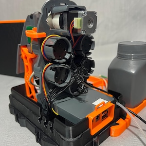 Peut inclure: Un dispositif m&eacute;canique complexe avec des composants gris et orange, comprenant des moteurs, des fils et une base noire. Une bouteille grise est visible &agrave; droite. L'appareil semble &ecirc;tre un prototype ou une pi&egrave;ce de machinerie.