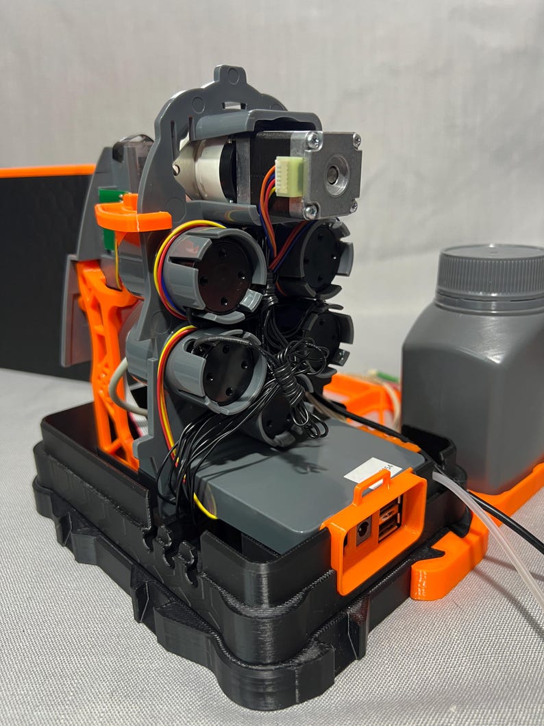Peut inclure: Un dispositif m&eacute;canique complexe avec des composants gris et orange, comprenant des moteurs, des fils et une base noire. Une bouteille grise est visible &agrave; droite. L'appareil semble &ecirc;tre un prototype ou une pi&egrave;ce de machinerie.