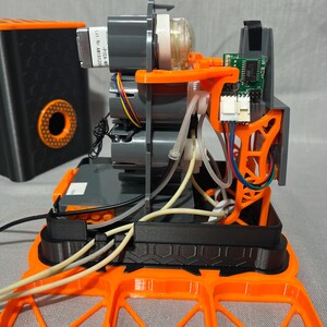 Peut inclure: Vue ouverte d'un appareil m&eacute;canique avec des moteurs gris, des supports orange et une carte de circuit imprim&eacute;. L'appareil comporte des tubes transparents et du c&acirc;blage. Un conteneur noir avec un design circulaire orange est &agrave; l'arri&egrave;re-plan.
