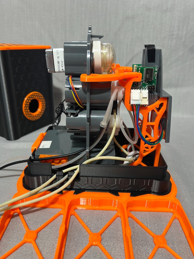 Peut inclure: Vue ouverte d'un appareil m&eacute;canique avec des moteurs gris, des supports orange et une carte de circuit imprim&eacute;. L'appareil comporte des tubes transparents et du c&acirc;blage. Un conteneur noir avec un design circulaire orange est &agrave; l'arri&egrave;re-plan.