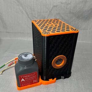 Peut inclure: Appareil rectangulaire noir et orange avec un motif en nid d'abeille sur le dessus et un motif circulaire sur le c&ocirc;t&eacute;. Une bouteille grise avec le texte "TRIDENT REAGENT ALKALINITY" est pos&eacute;e sur une base orange &agrave; c&ocirc;t&eacute; de l'appareil. L'appareil a une s&eacute;rie de fils attach&eacute;s.