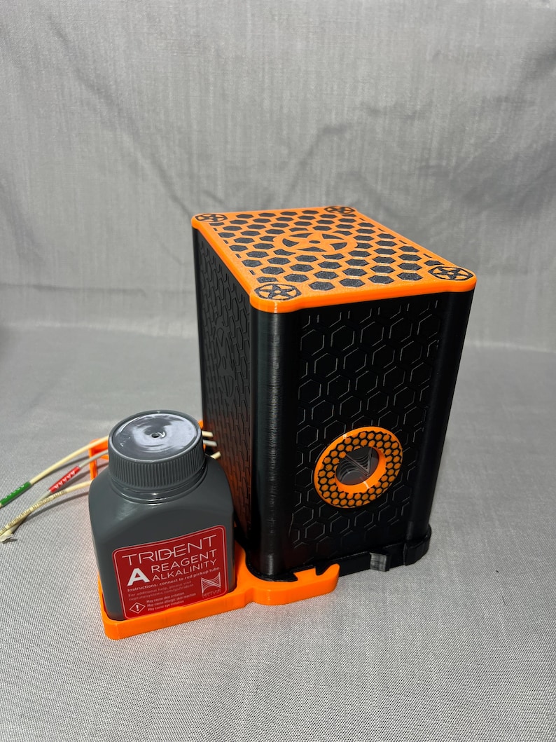Peut inclure: Appareil rectangulaire noir et orange avec un motif en nid d'abeille sur le dessus et un motif circulaire sur le c&ocirc;t&eacute;. Une bouteille grise avec le texte "TRIDENT REAGENT ALKALINITY" est pos&eacute;e sur une base orange &agrave; c&ocirc;t&eacute; de l'appareil. L'appareil a une s&eacute;rie de fils attach&eacute;s.