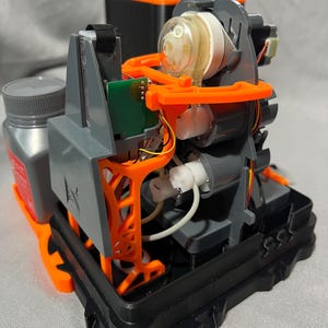 Peut inclure: Un appareil d&eacute;mont&eacute; r&eacute;v&eacute;lant sa structure interne complexe. Il est compos&eacute; de pi&egrave;ces grises, noires et oranges, dont un &eacute;l&eacute;ment cylindrique transparent, une petite carte de circuit imprim&eacute; verte et un flacon gris avec un bouchon gris. Fond gris clair.