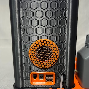 Peut inclure: Un appareil noir et orange imprim&eacute; en 3D avec un motif en nid d'abeille. L'appareil poss&egrave;de un haut-parleur orange circulaire et un panneau orange avec des ports. Le dessus et la base sont &eacute;galement orange.