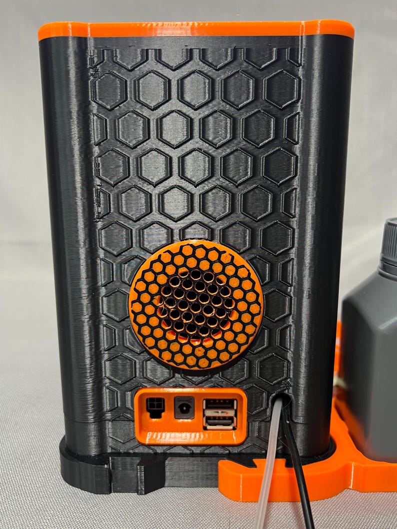 Peut inclure: Un appareil noir et orange imprim&eacute; en 3D avec un motif en nid d'abeille. L'appareil poss&egrave;de un haut-parleur orange circulaire et un panneau orange avec des ports. Le dessus et la base sont &eacute;galement orange.
