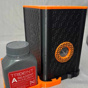 Peut inclure: Un kit de test de r&eacute;actif d'alcalinit&eacute; Trident noir et orange. Le kit comprend une bouteille grise avec du texte rouge et blanc, et un conteneur noir avec un motif en nid d'abeille. La bouteille indique "TRIDENT REAGENT ALKALINITY".