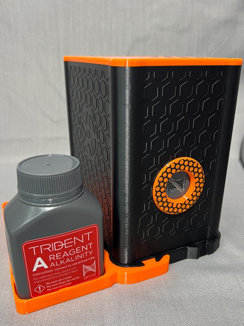 Peut inclure: Un kit de test de r&eacute;actif d'alcalinit&eacute; Trident noir et orange. Le kit comprend une bouteille grise avec du texte rouge et blanc, et un conteneur noir avec un motif en nid d'abeille. La bouteille indique "TRIDENT REAGENT ALKALINITY".