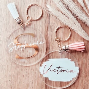 Puede incluir: Dos llaveros redondos de acrílico transparente con herrajes dorados y borlas. Un llavero presenta el nombre "Stephanie" con una inicial dorada. El otro llavero tiene el nombre "Victoria" sobre un fondo azul claro.