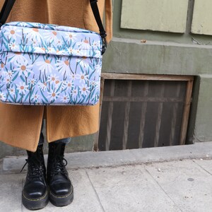 May include: A blue and white floral print messenger bag with a black strap. The bag is being held by a person wearing a brown coat and black boots.