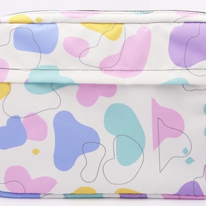May include: A white crossbody bag with a zipper closure and a colorful abstract pattern of pink, yellow, blue, and purple shapes. The bag has a black adjustable strap.