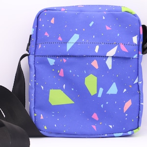 May include: A blue crossbody bag with a black adjustable strap. The bag has a colorful confetti pattern with pink, green, blue, and white shapes.