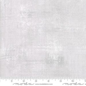 May include: A close-up of a light gray fabric with a subtle textured pattern. The fabric has a slightly distressed look, with a hint of white throughout. The fabric is marked with a ruler, showing the measurements in inches.